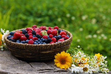 Wicker bowl with different fresh ripe berries and beautiful flowers on wooden surface outdoors