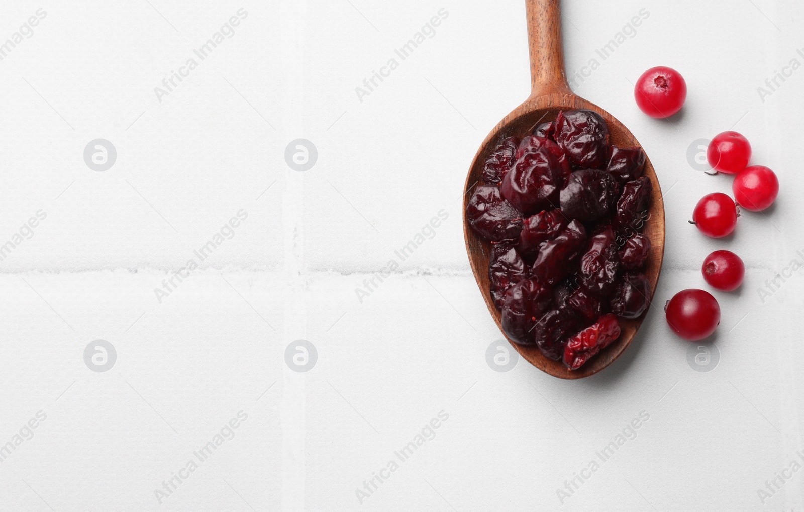Photo of Tasty dried cranberries in spoon and fresh ones on white tiled table, top view. Space for text