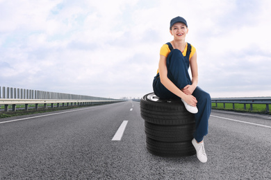 Mechanic in uniform with car tires on asphalt highway outdoors, space for text