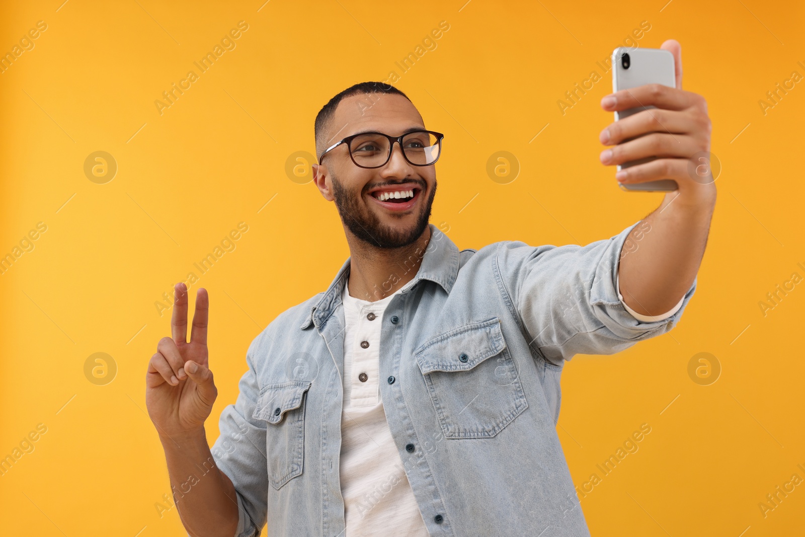 Photo of Smiling young man taking selfie with smartphone and showing peace sign on yellow background