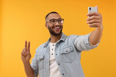 Photo of Smiling young man taking selfie with smartphone and showing peace sign on yellow background