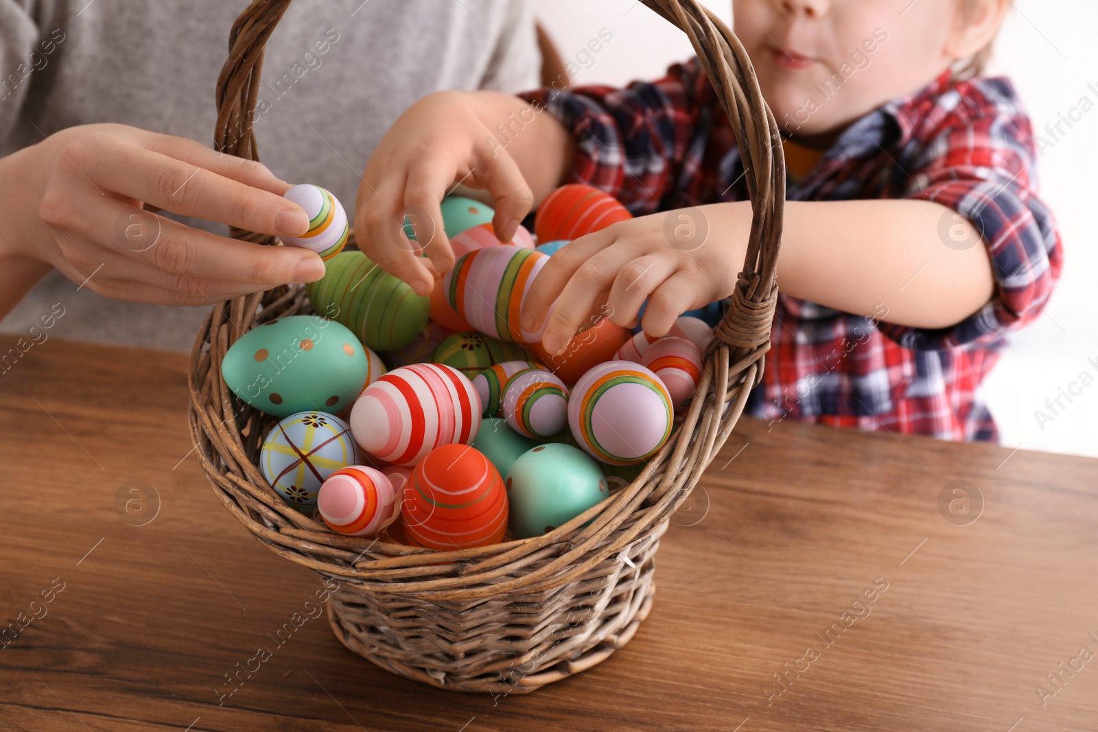 Photo of Little boy and his mother with basket full of dyed Easter eggs at wooden table, closeup. Easter celebration