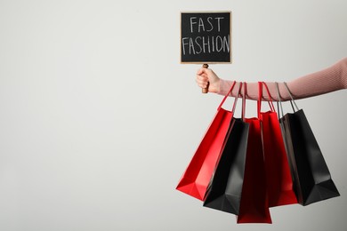 Woman holding small chalkboard with phrase Fast Fashion and different shopping bags on light background, closeup. Space for text