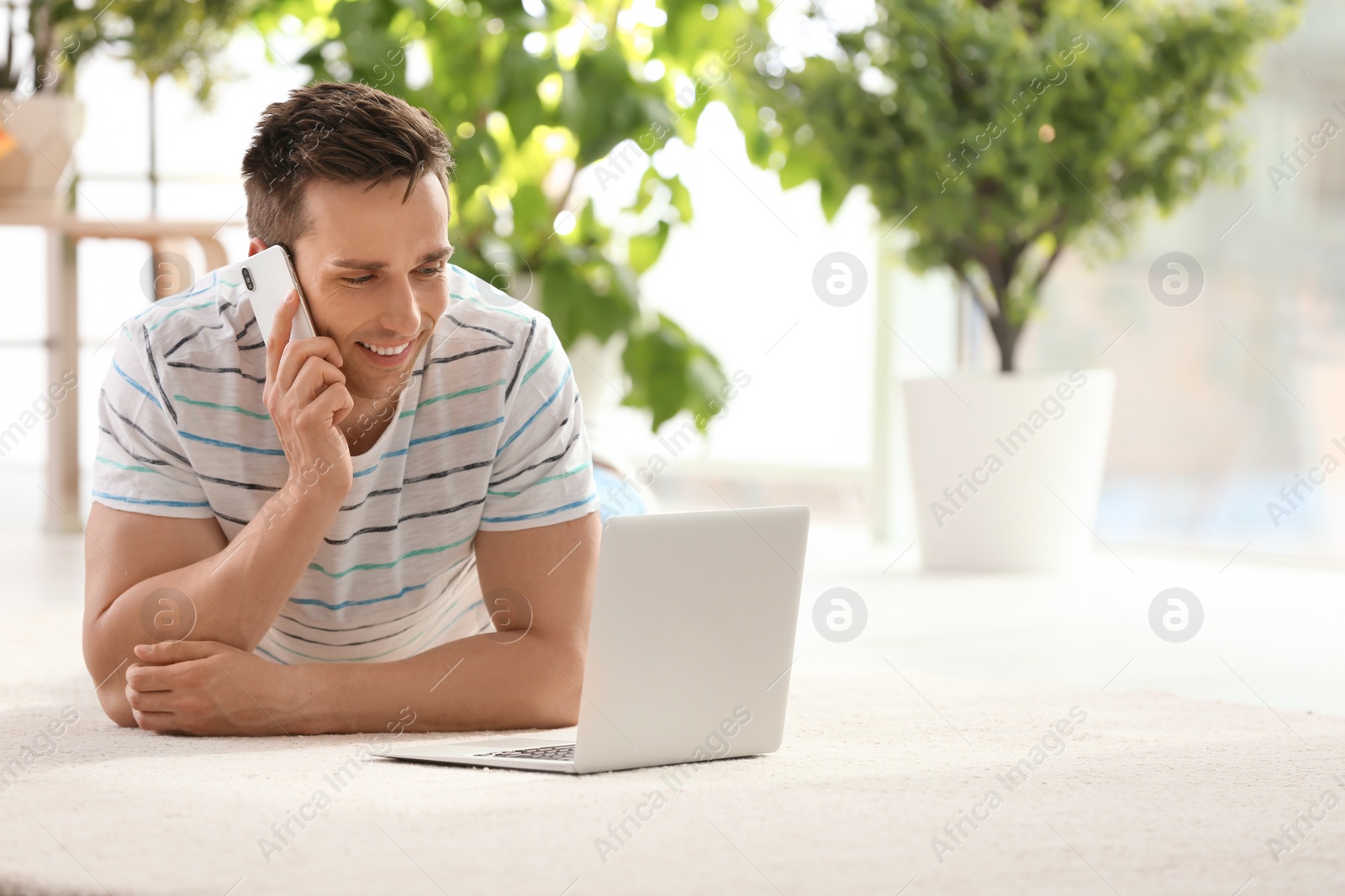 Photo of Handsome young man with mobile phone and laptop on cozy carpet at home