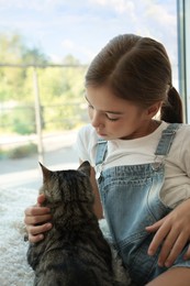 Cute little girl with her cat near window at home. Childhood pet