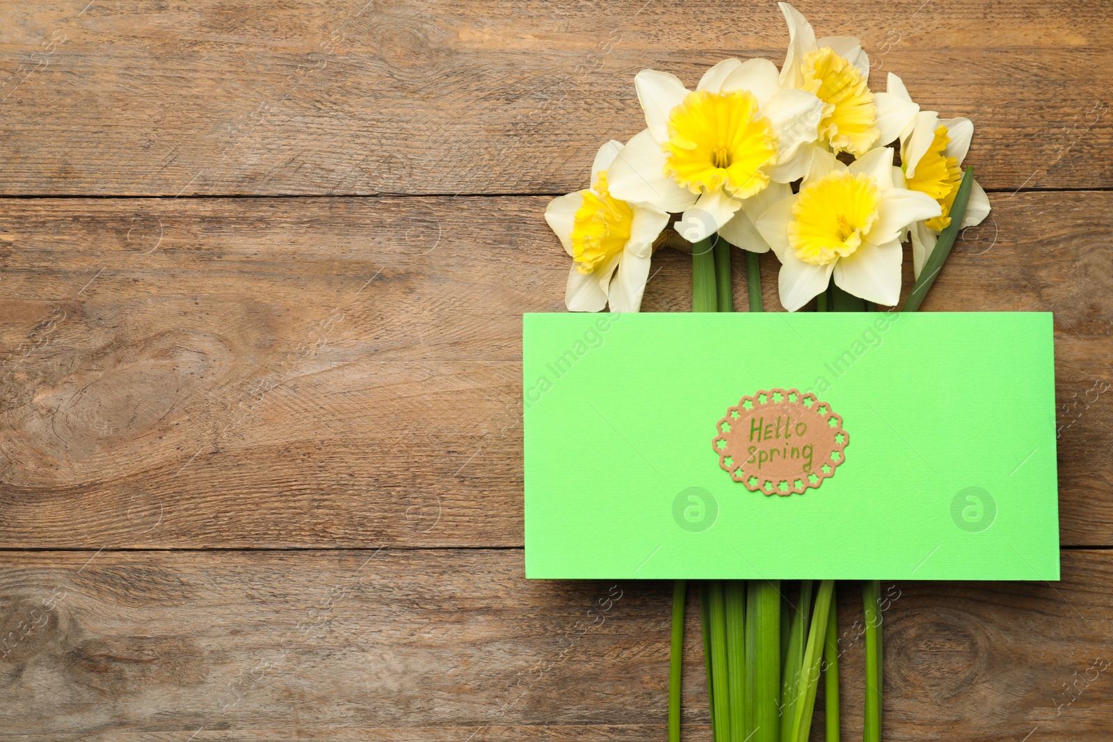 Photo of Green card with words HELLO SPRING and narcissus flowers on wooden table, flat lay. Space for text