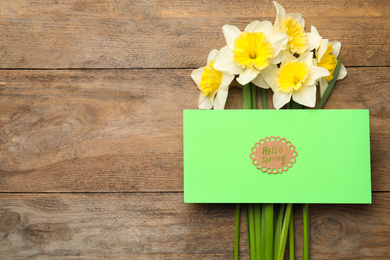 Photo of Green card with words HELLO SPRING and narcissus flowers on wooden table, flat lay. Space for text