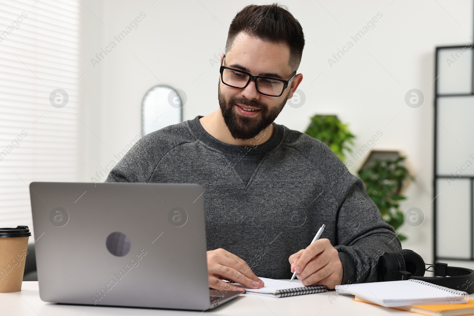 Photo of E-learning. Young man taking notes during online lesson at white table indoors