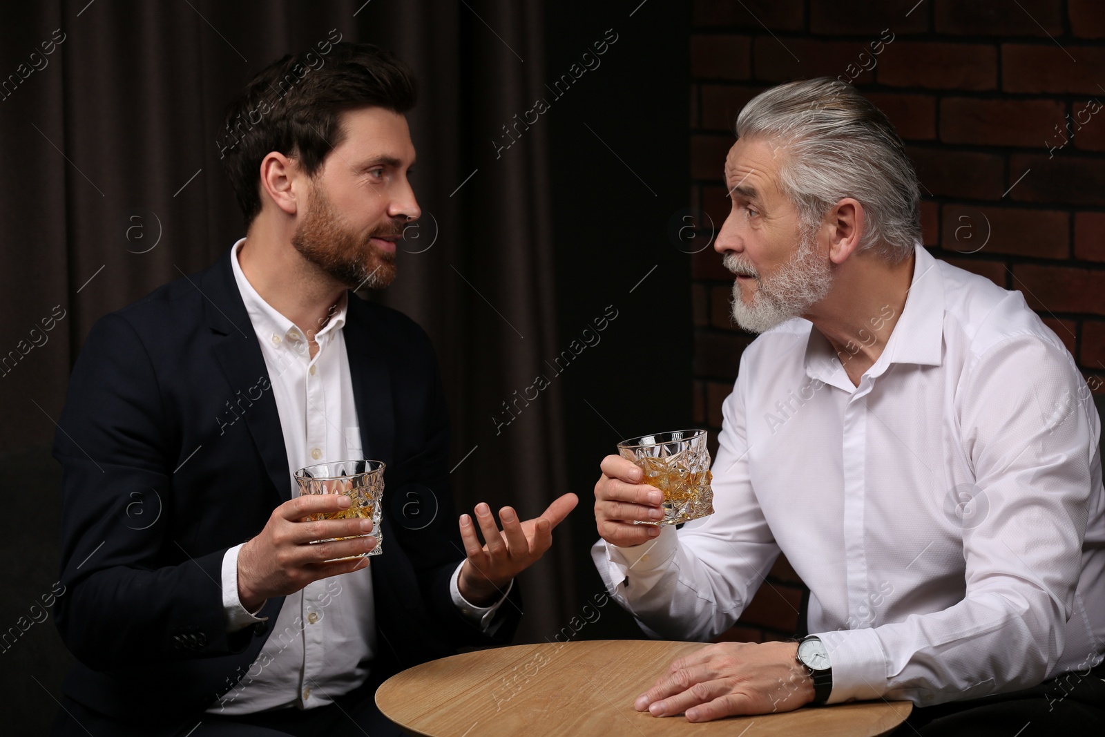 Photo of Men with glasses of whiskey talking at wooden table indoors