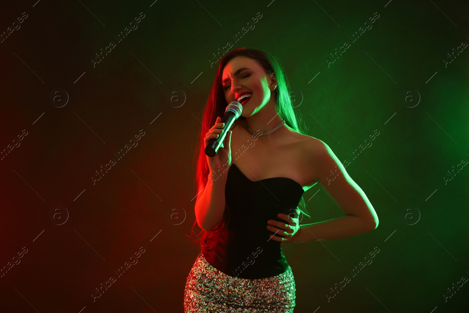 Photo of Beautiful woman with microphone singing in color lights on dark background. Space for text