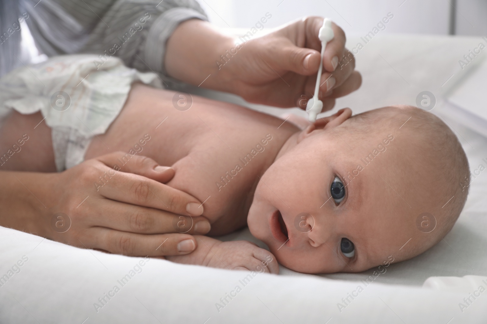 Photo of Mother cleaning ears of her baby with cotton bud on changing table indoors, closeup