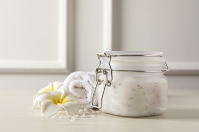 Body scrub in glass jar, towel and plumeria flowers on white wooden table