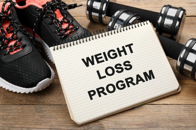 Photo of Notebook with phrase Weight Loss Program, sneakers and dumbbells on wooden table