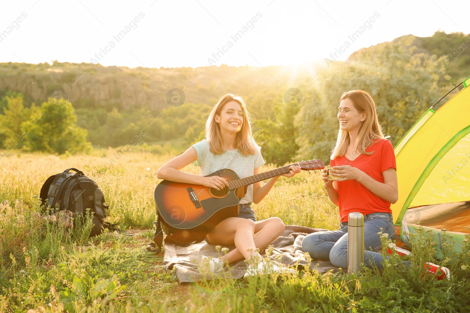 Photo of Young women resting with hot drink and guitar near camping tent in wilderness