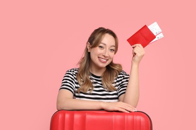 Happy young woman with passport, ticket and suitcase on pink background