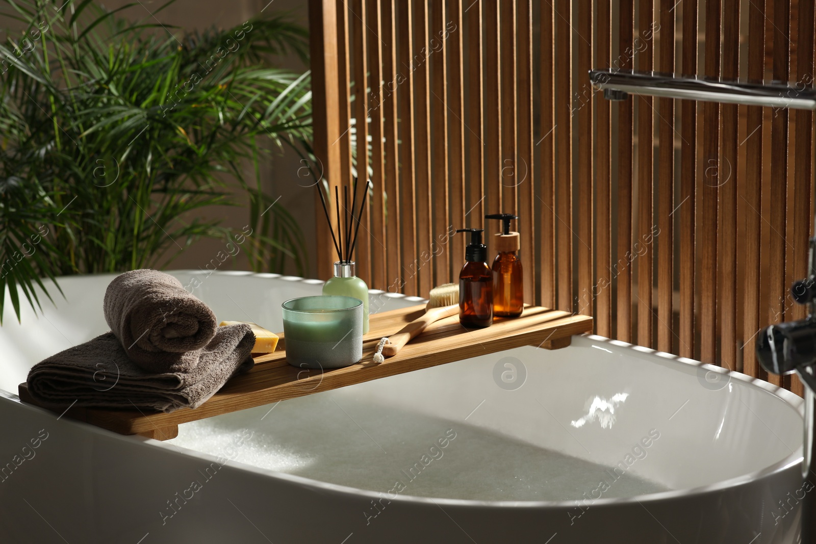 Photo of Wooden bath tray with candle, air freshener and bathroom amenities on tub indoors