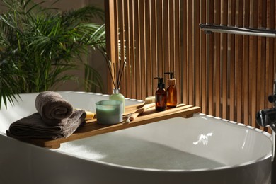 Photo of Wooden bath tray with candle, air freshener and bathroom amenities on tub indoors
