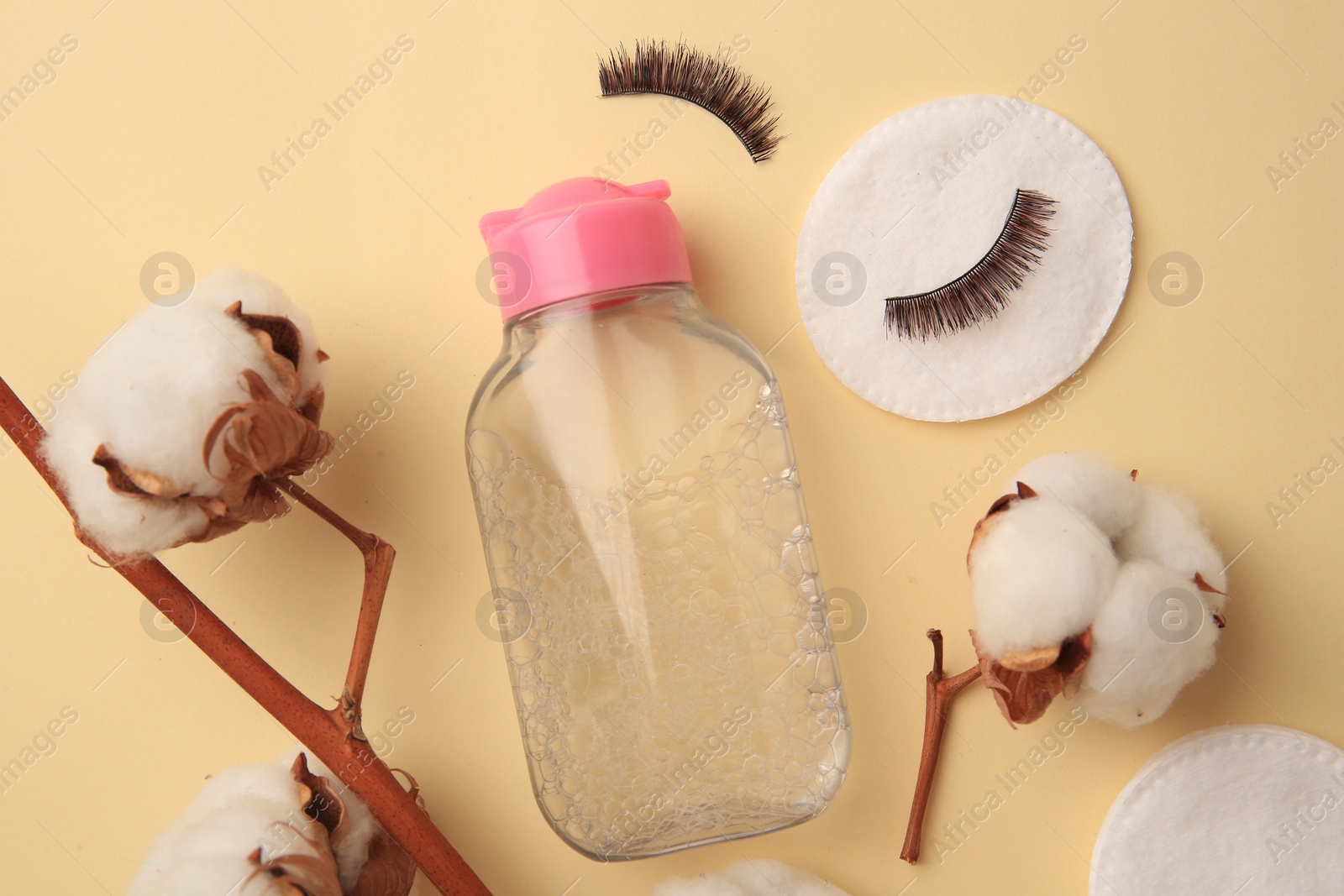 Photo of Bottle of makeup remover, cotton flowers, pads and false eyelashes on yellow background, flat lay
