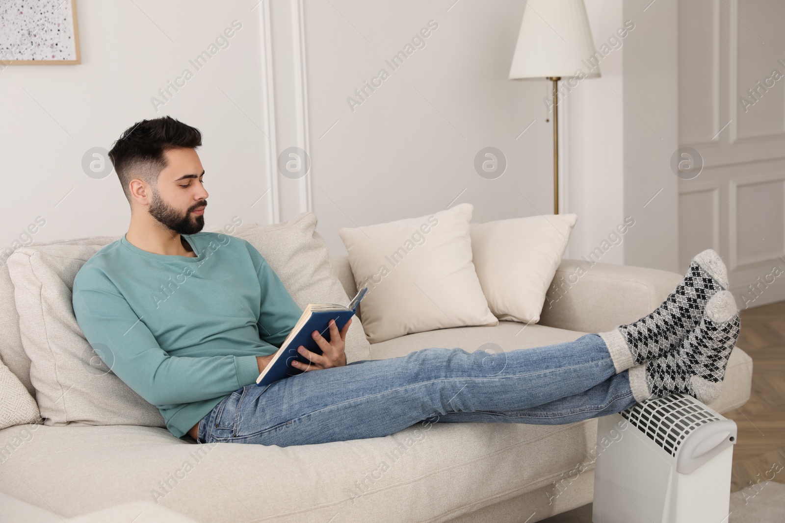 Photo of Young man warming feet on electric heater at home