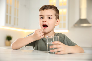 Little boy with glass of water taking vitamin capsule in kitchen