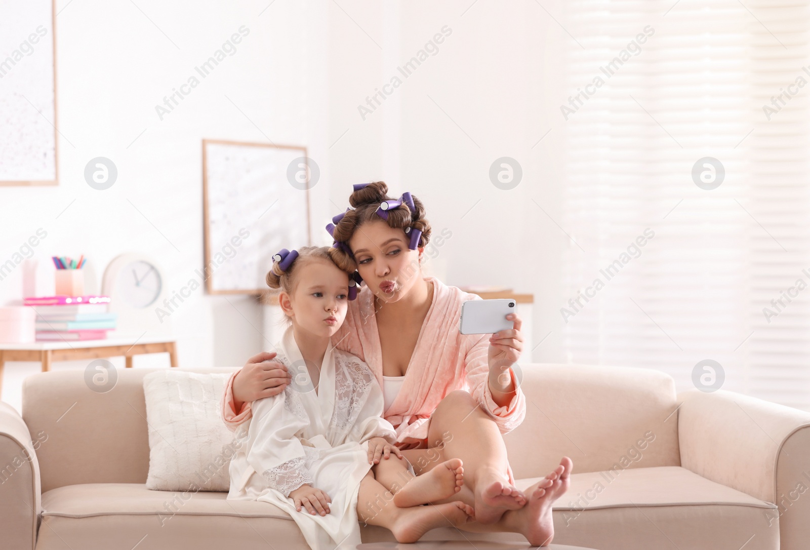 Photo of Happy mother and daughter with curlers and mobile phone on sofa at home