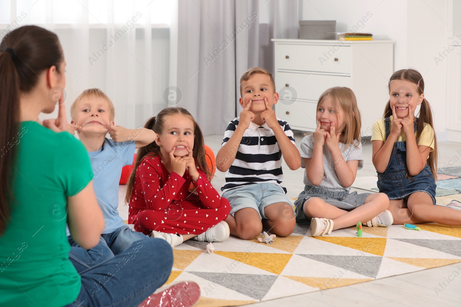 Photo of Nursery teacher and group of cute little children studying with fun on bright rug in kindergarten. Playtime activities