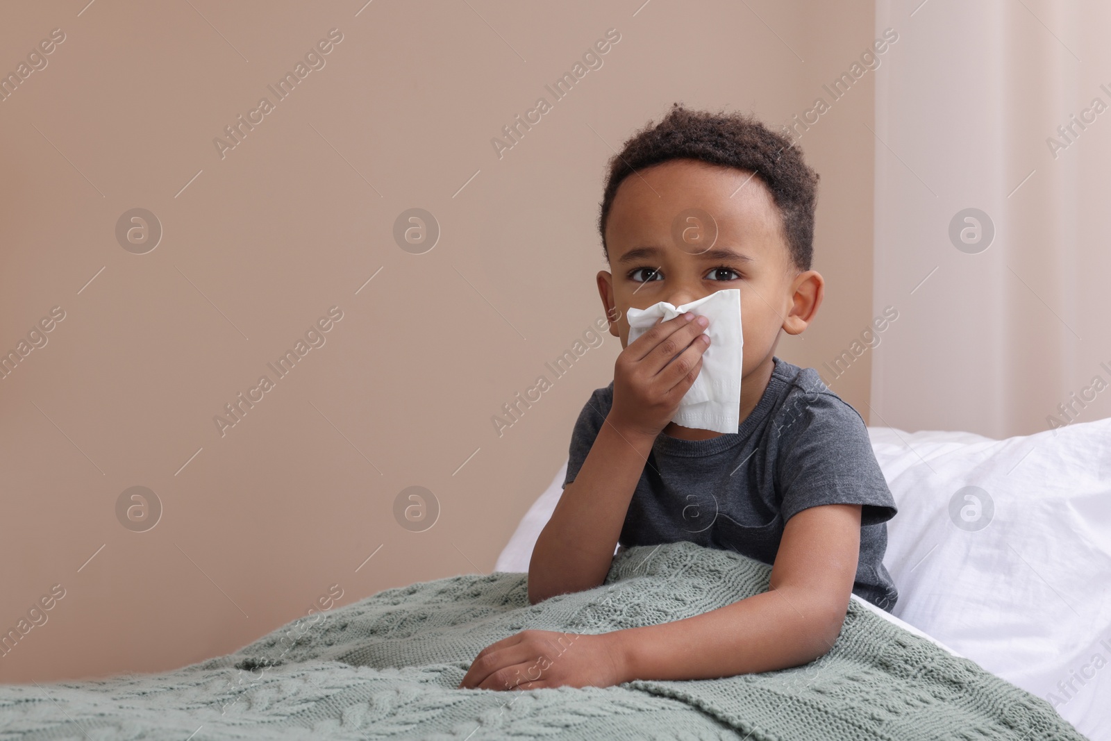 Photo of African-American boy with tissue blowing nose in bed indoors, space for text. Cold symptoms