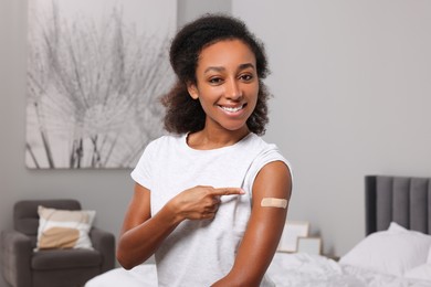 Photo of Happy young woman pointing at adhesive bandage after vaccination indoors