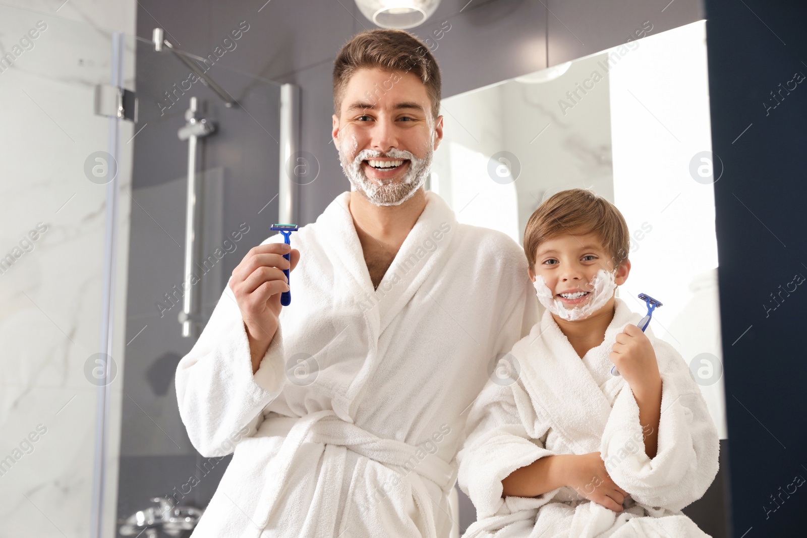 Photo of Dad and son with shaving foam on faces holding razors in bathroom