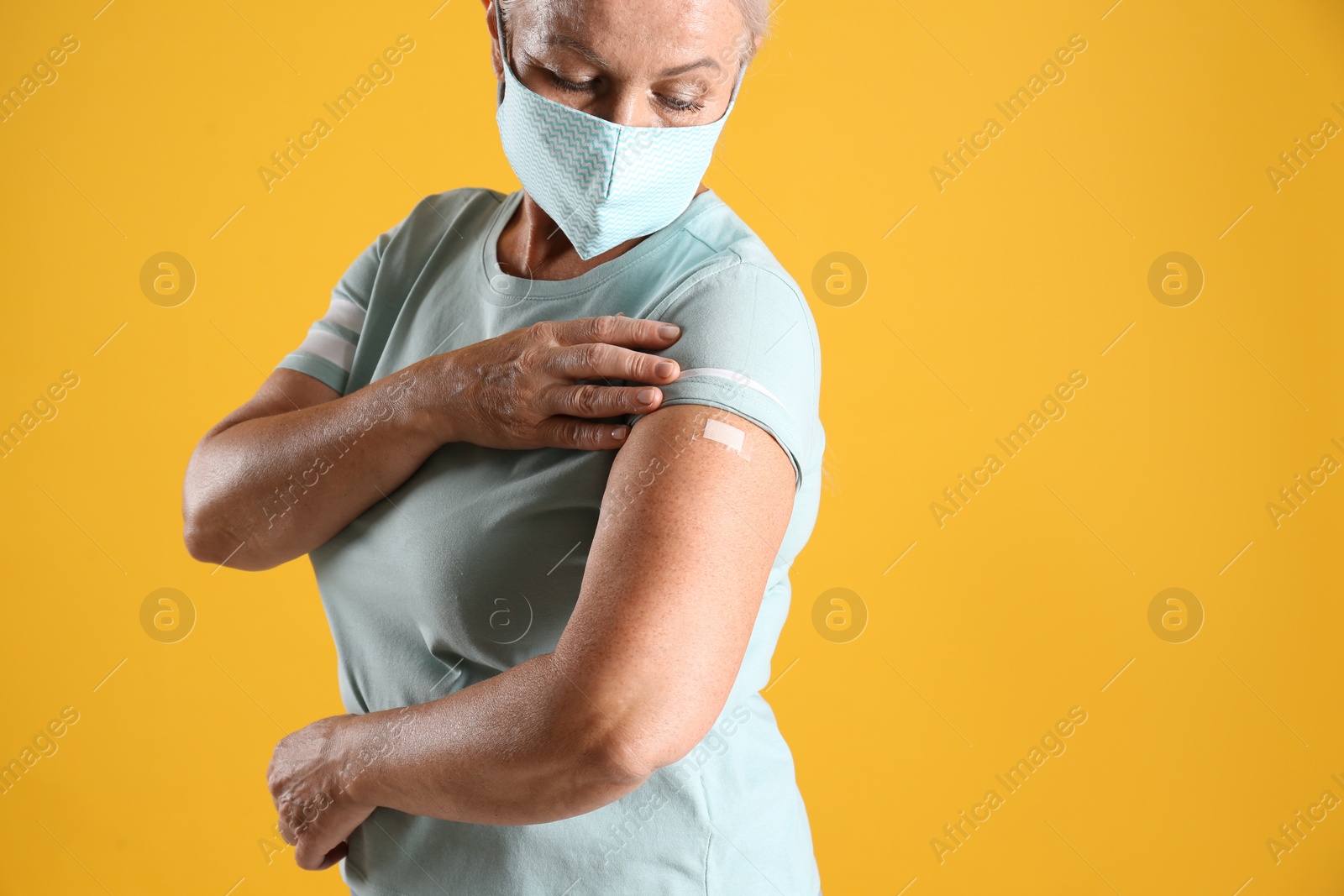 Photo of Mature woman in protective mask showing arm with bandage after vaccination on yellow background. Space for text