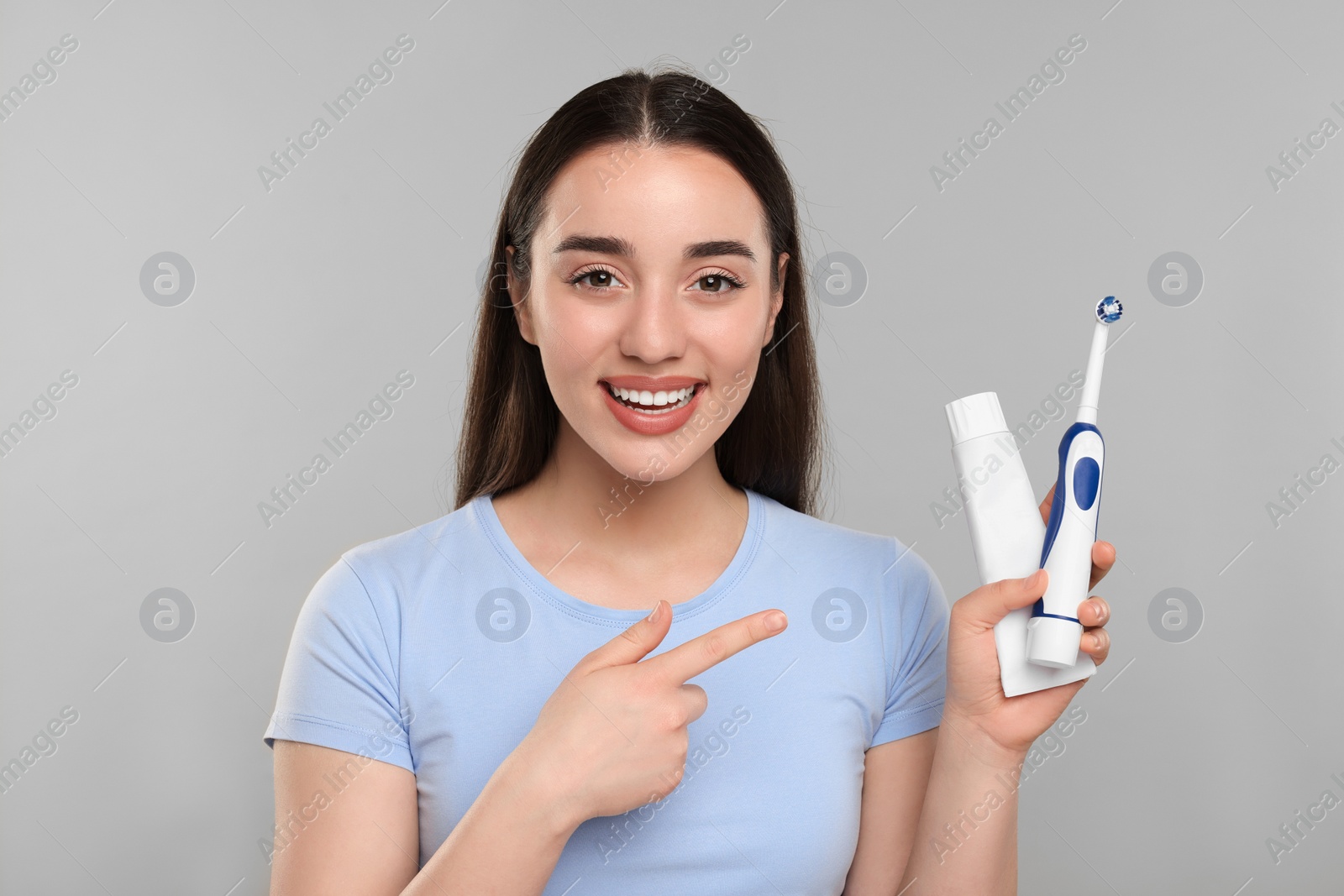 Photo of Happy young woman holding electric toothbrush and tube of toothpaste on light grey background