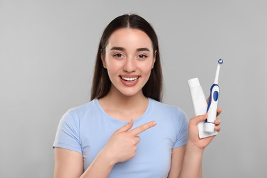 Happy young woman holding electric toothbrush and tube of toothpaste on light grey background