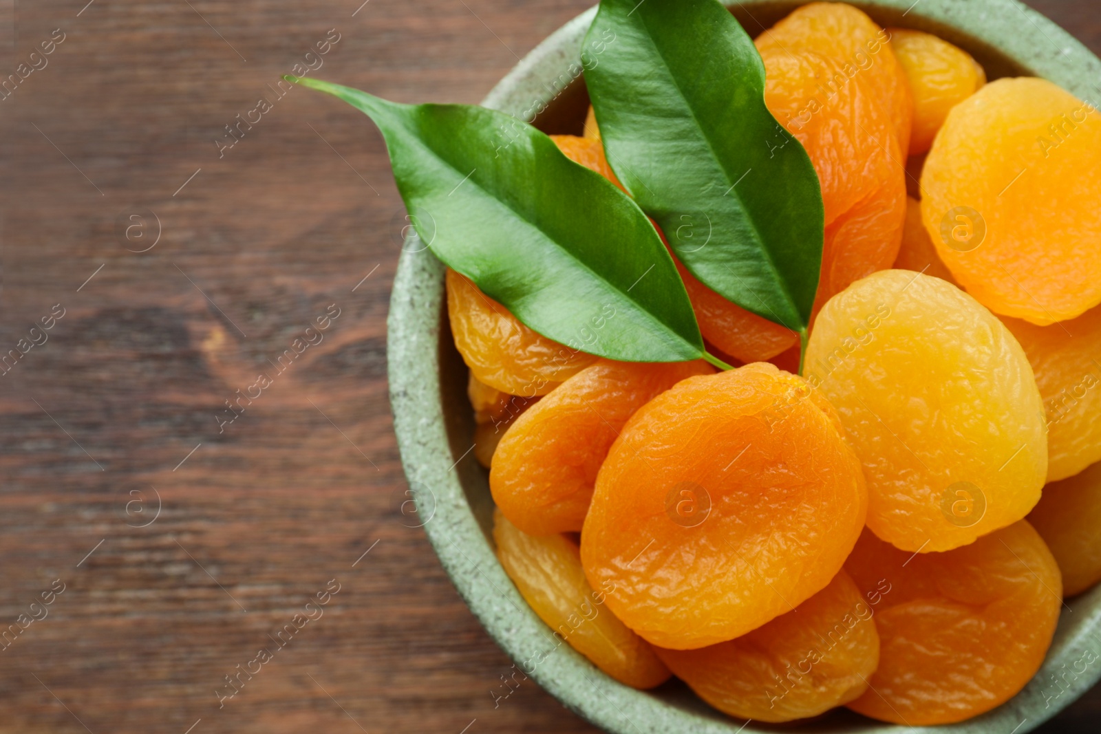 Photo of Bowl of tasty apricots and green leaves on wooden table, top view with space for text. Dried fruits