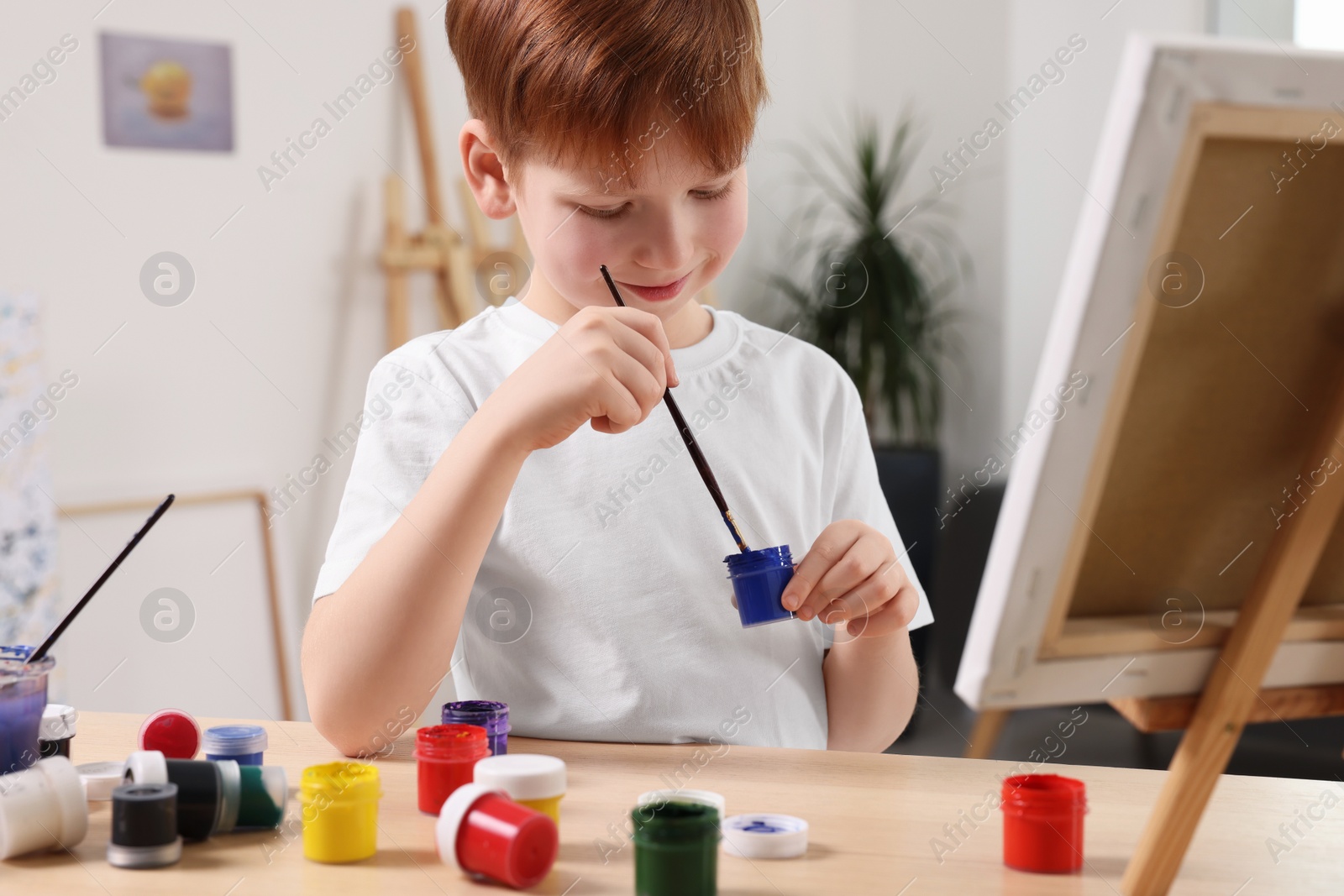 Photo of Little boy painting at table in studio. Using easel to hold canvas