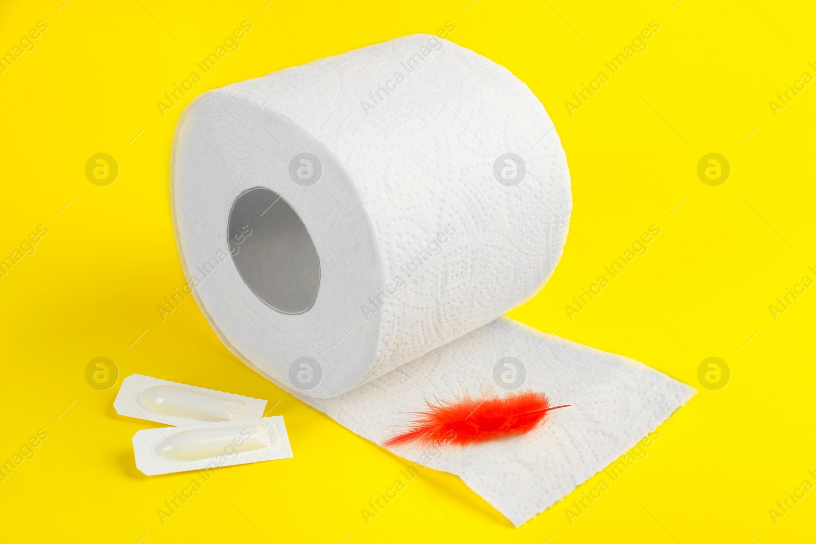 Photo of Roll of toilet paper, red feather and suppositories on yellow background. Hemorrhoid problems