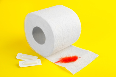 Photo of Roll of toilet paper, red feather and suppositories on yellow background. Hemorrhoid problems