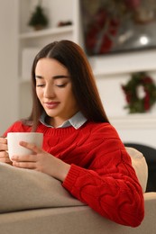 Photo of Woman holding cup of hot drink on sofa near Christmas tree indoors