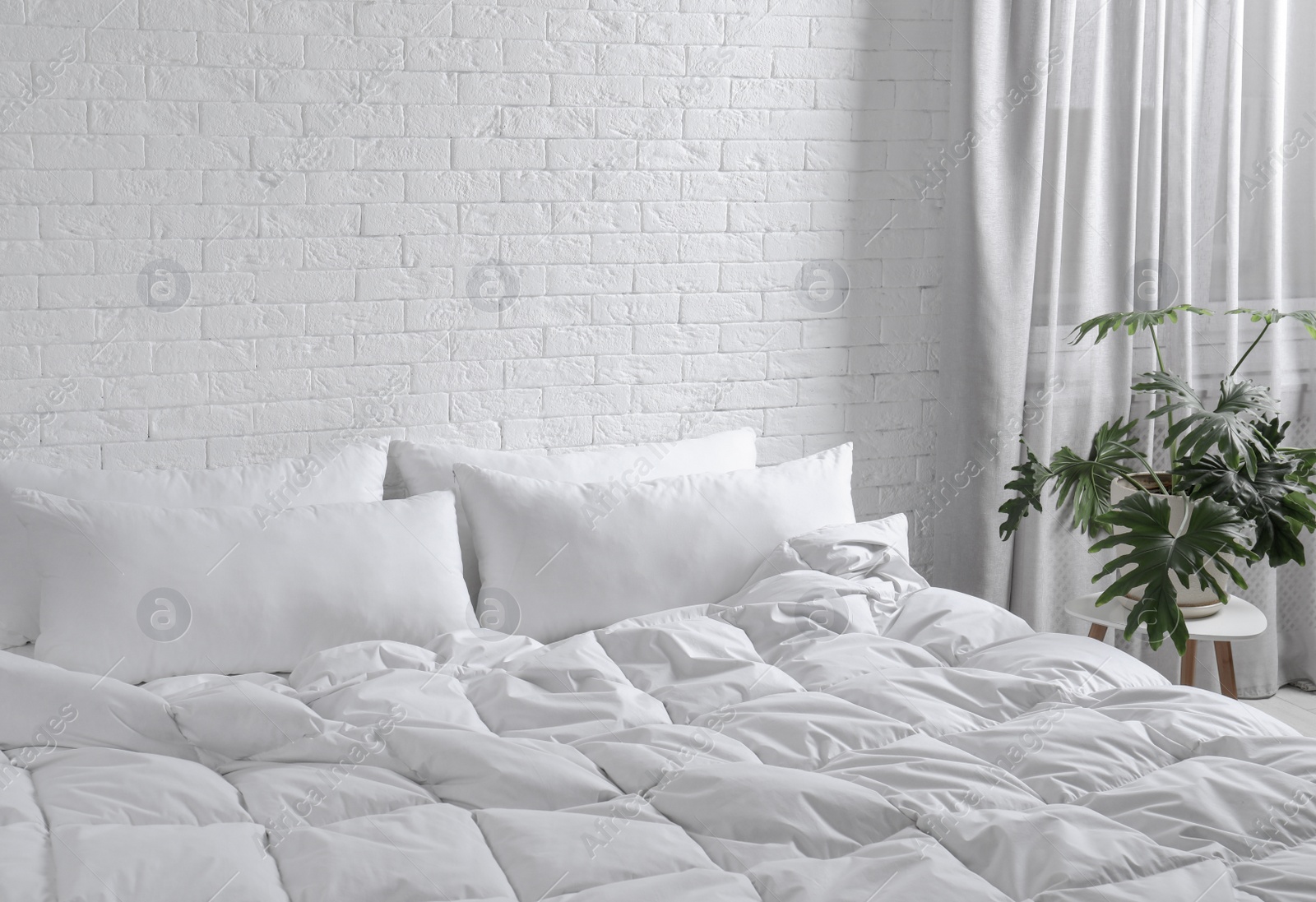 Photo of Large comfortable bed with pillows and blanket near white brick wall indoors. Stylish interior