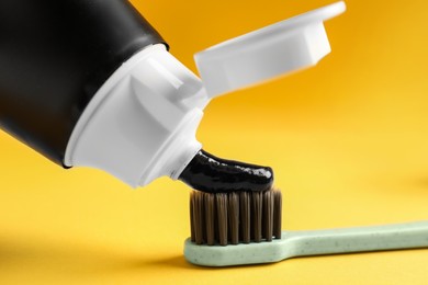 Photo of Applying charcoal toothpaste on brush against yellow background, closeup