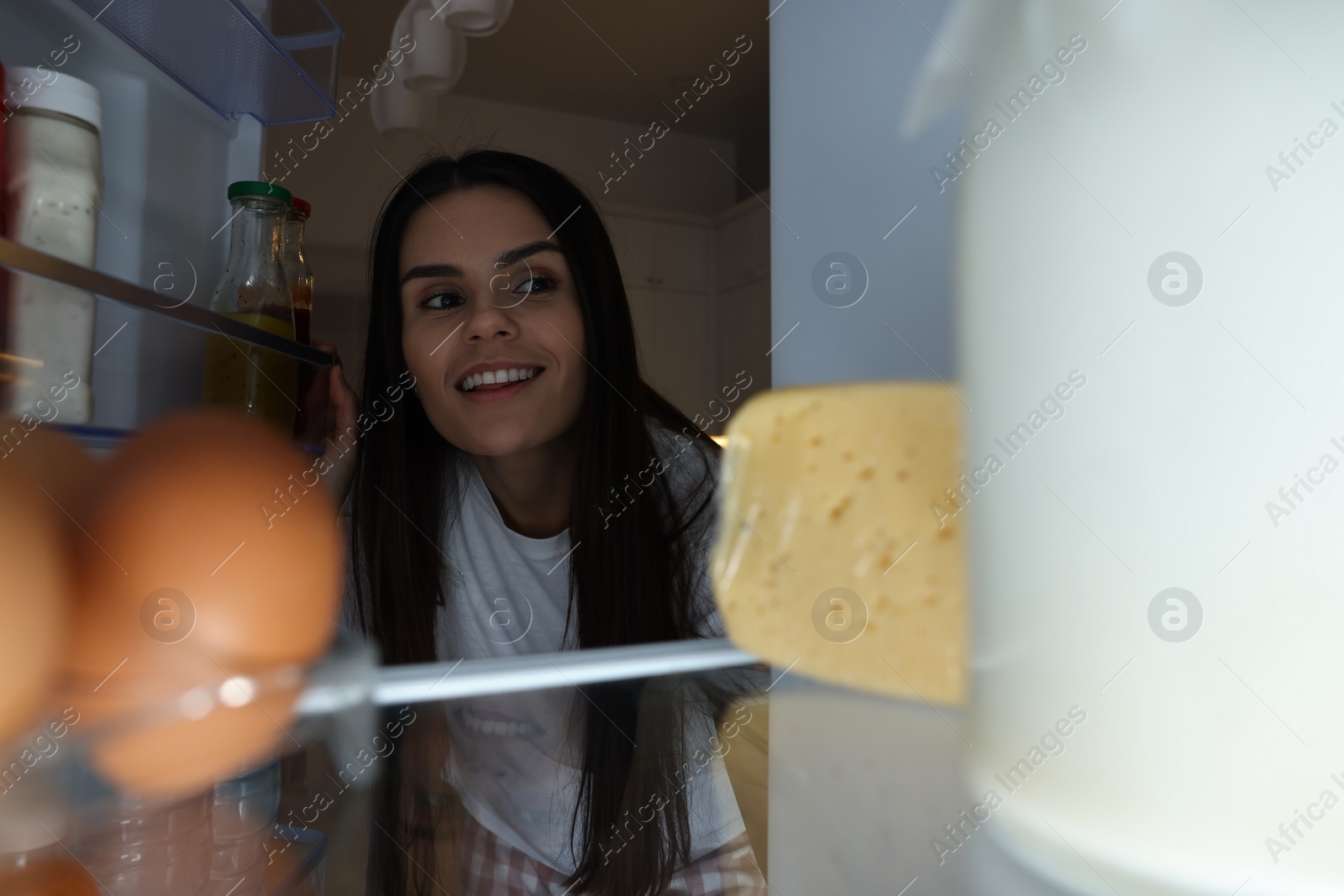 Photo of Young woman near modern refrigerator in kitchen at night, view from inside