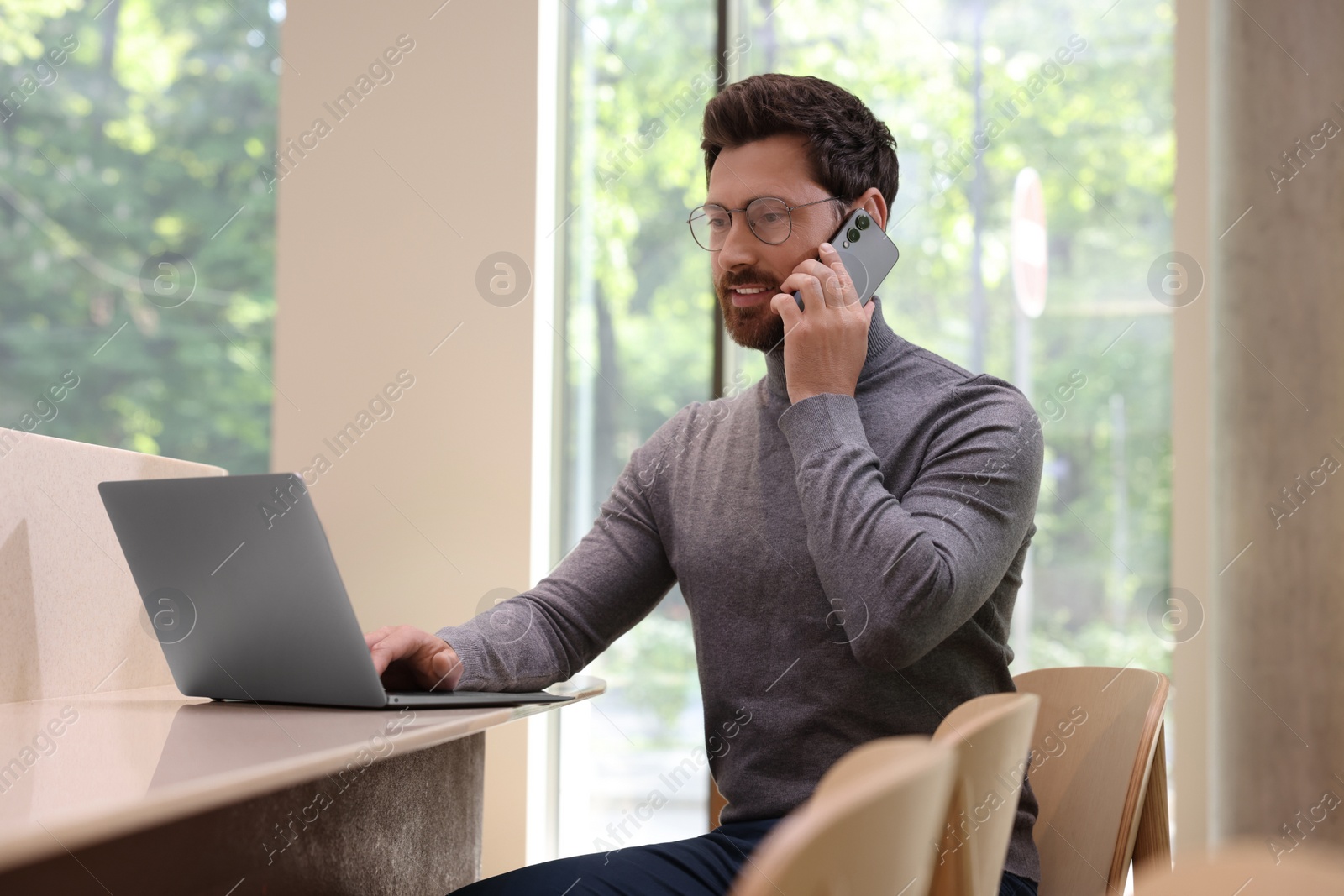 Photo of Handsome man talking on smartphone while using laptop at table in cafe