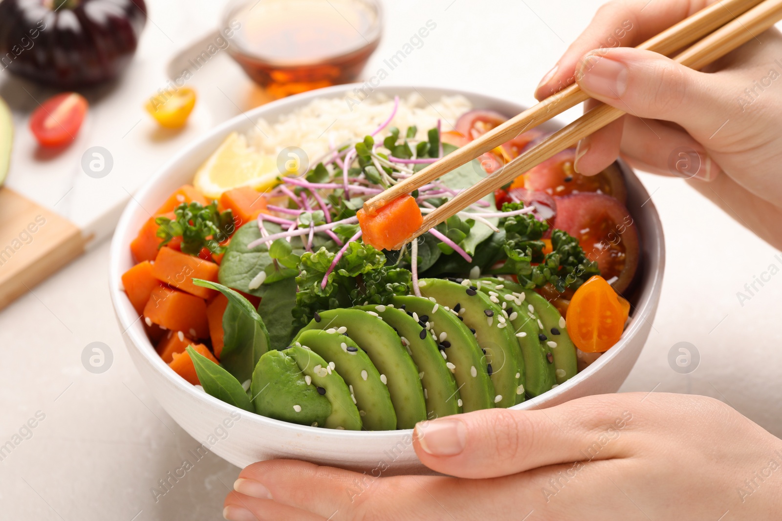 Photo of Woman eating delicious vegan bowl with avocados, carrots and tomatoes at light table, closeup