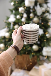 Woman with cup of delicious hot drink near Christmas tree at home, closeup