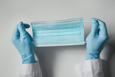 Photo of Doctor in latex gloves holding disposable face mask on light background, closeup. Protective measures during coronavirus quarantine