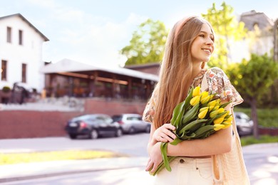 Beautiful teenage girl with bouquet of yellow tulips on city street, space for text