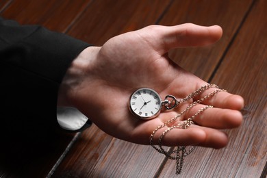 Man holding chain with elegant pocket watch at wooden table, closeup