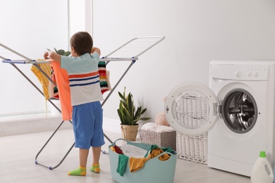 Photo of Cute little boy hanging laundry onto clothes drying rack indoors, back view