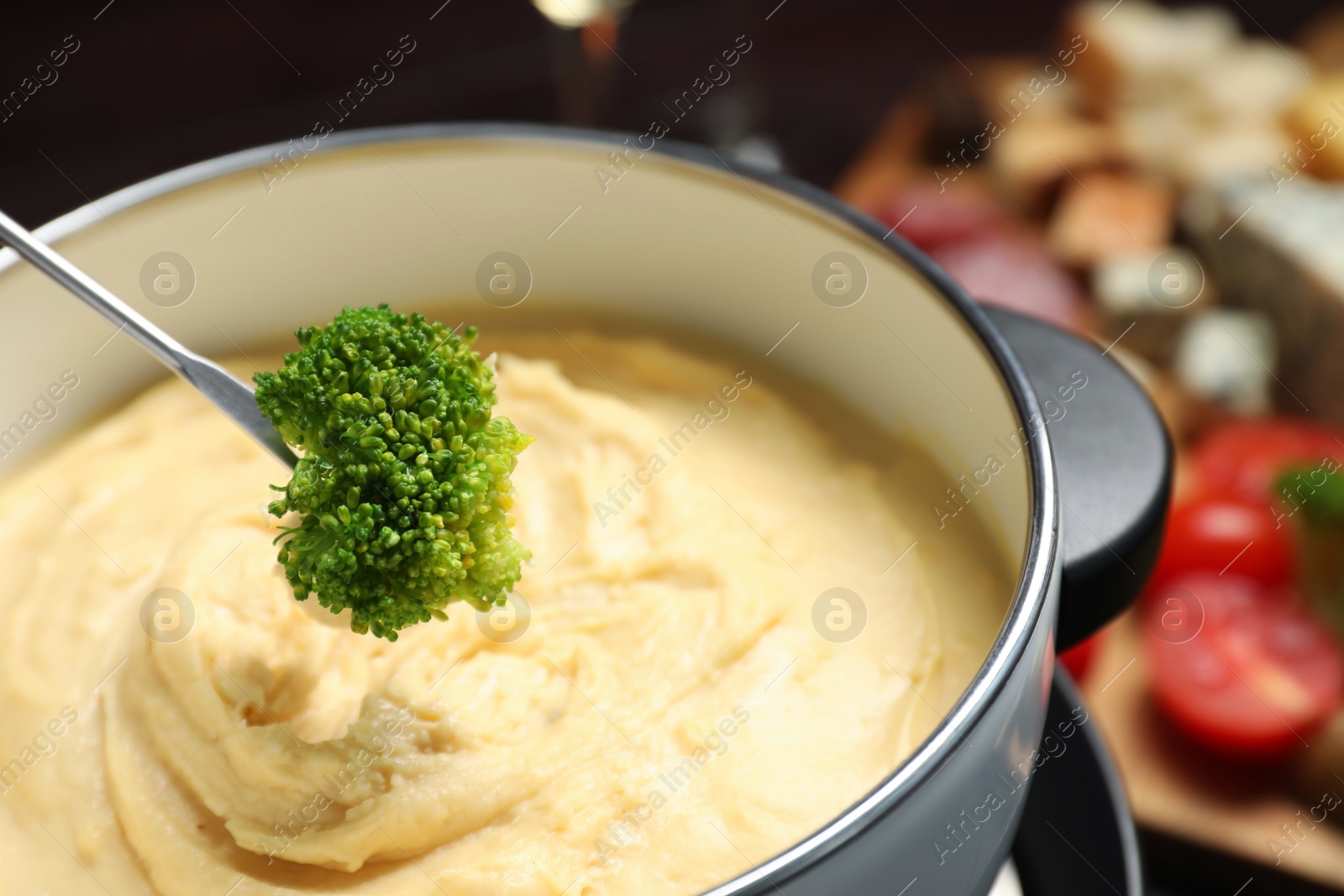 Photo of Dipping piece of broccoli into fondue pot with melted cheese on blurred background, closeup