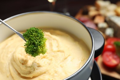 Photo of Dipping piece of broccoli into fondue pot with melted cheese on blurred background, closeup