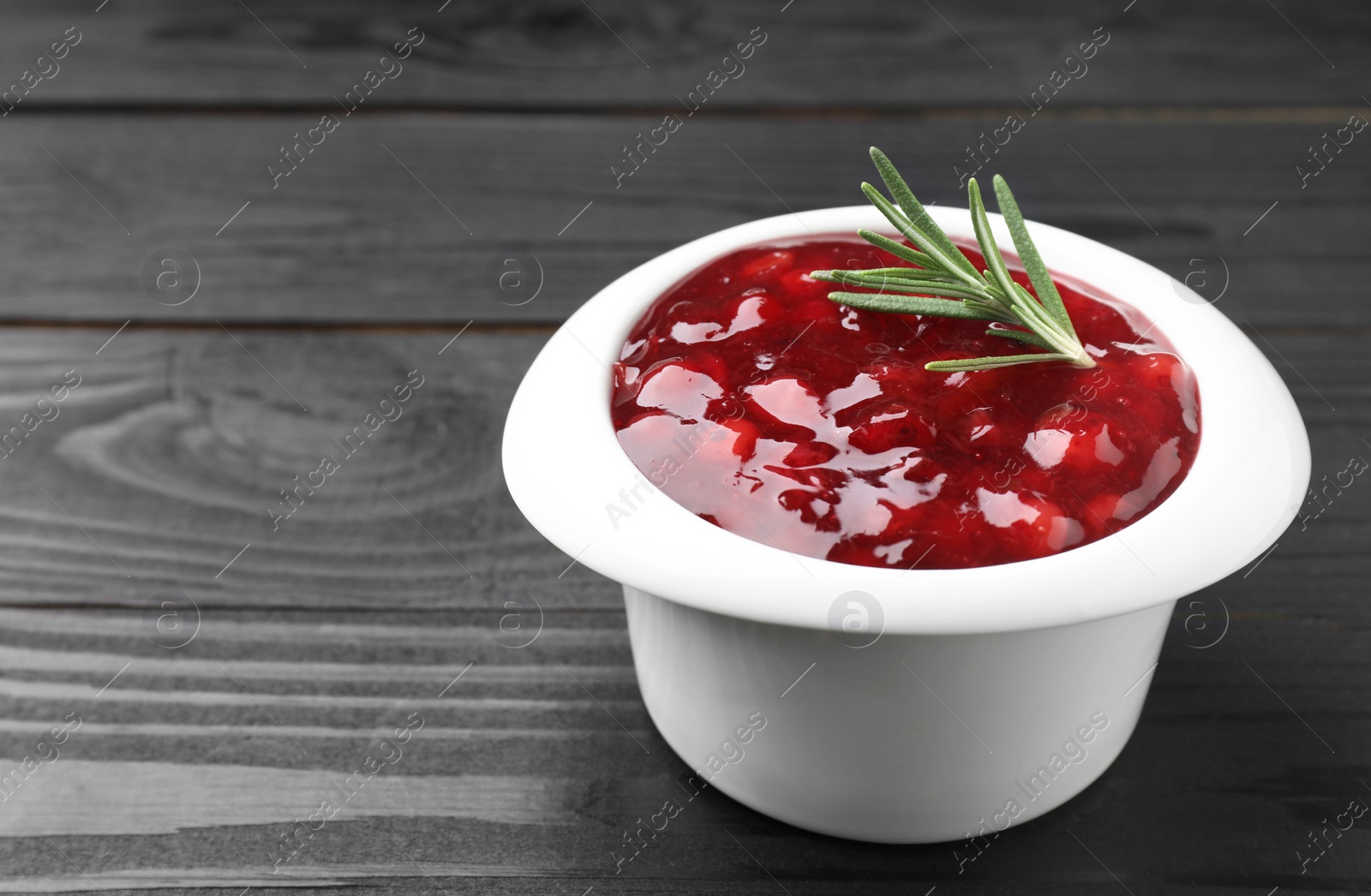 Photo of Fresh cranberry sauce in bowl and rosemary on black wooden table, closeup. Space for text